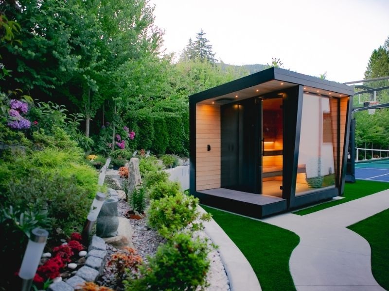 Modern outdoor sauna with black framing, warm wood accents and large glass panels positioned beside a curved pathway in a landscaped backyard with mature trees and garden beds.