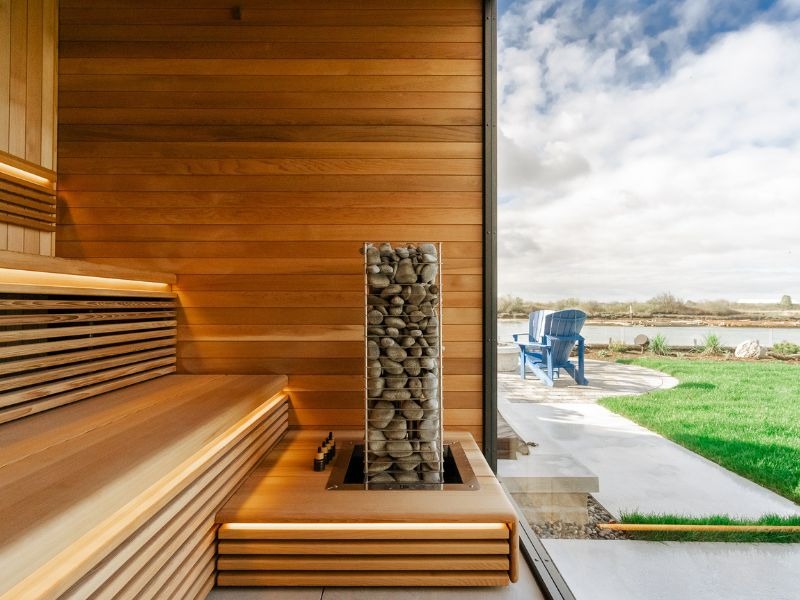Warm wood sauna interior with tiered benches, stone-filled electric heater and small sauna oil bottles positioned beside the heater, overlooking an outdoor waterfront setting.