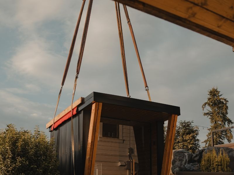 Modern outdoor sauna being lifted by crane straps during delivery and positioning at a residential property.