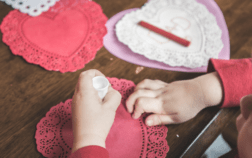 A toddler using sticky glue to attach heart-shaped paper cutouts, engaging in a sensory activity to develop fine motor skills.