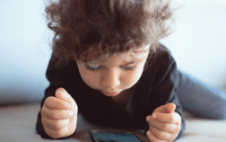 A toddler sitting on the floor, looking at a cellphone screen with a focused expression.