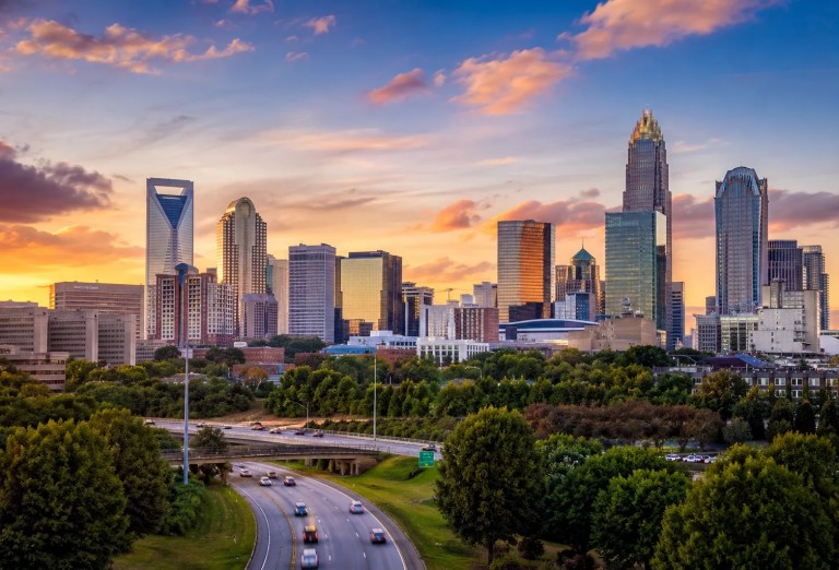Charlotte, North Carolina skyline at sunset with modern skyscrapers, tree-lined neighborhoods, and a highway leading into Uptown Charlotte.