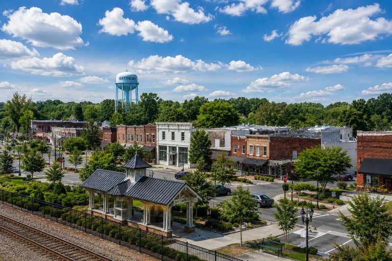 Historic downtown Matthews, North Carolina with the railroad tracks, pavilion, Main Street storefronts, and the Matthews water tower under a bright blue summer sky.