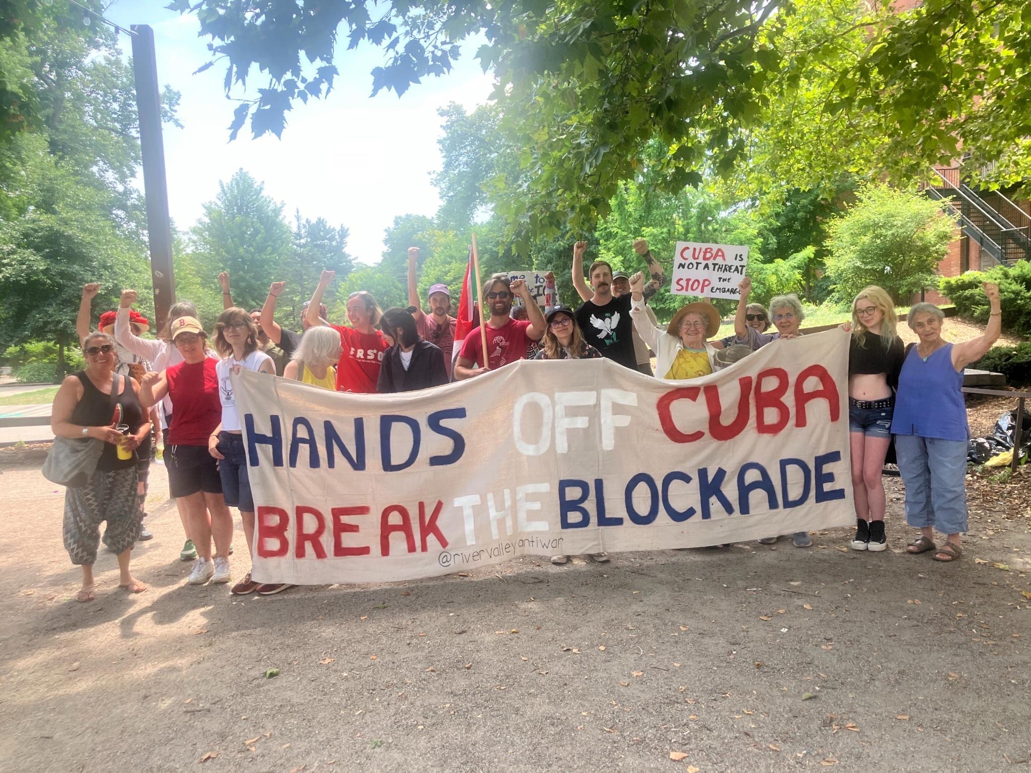 A group holds a wide canvas banner on a sunny day in Pulaski Park reading "Hands Off Cuba, Break the Blockade."