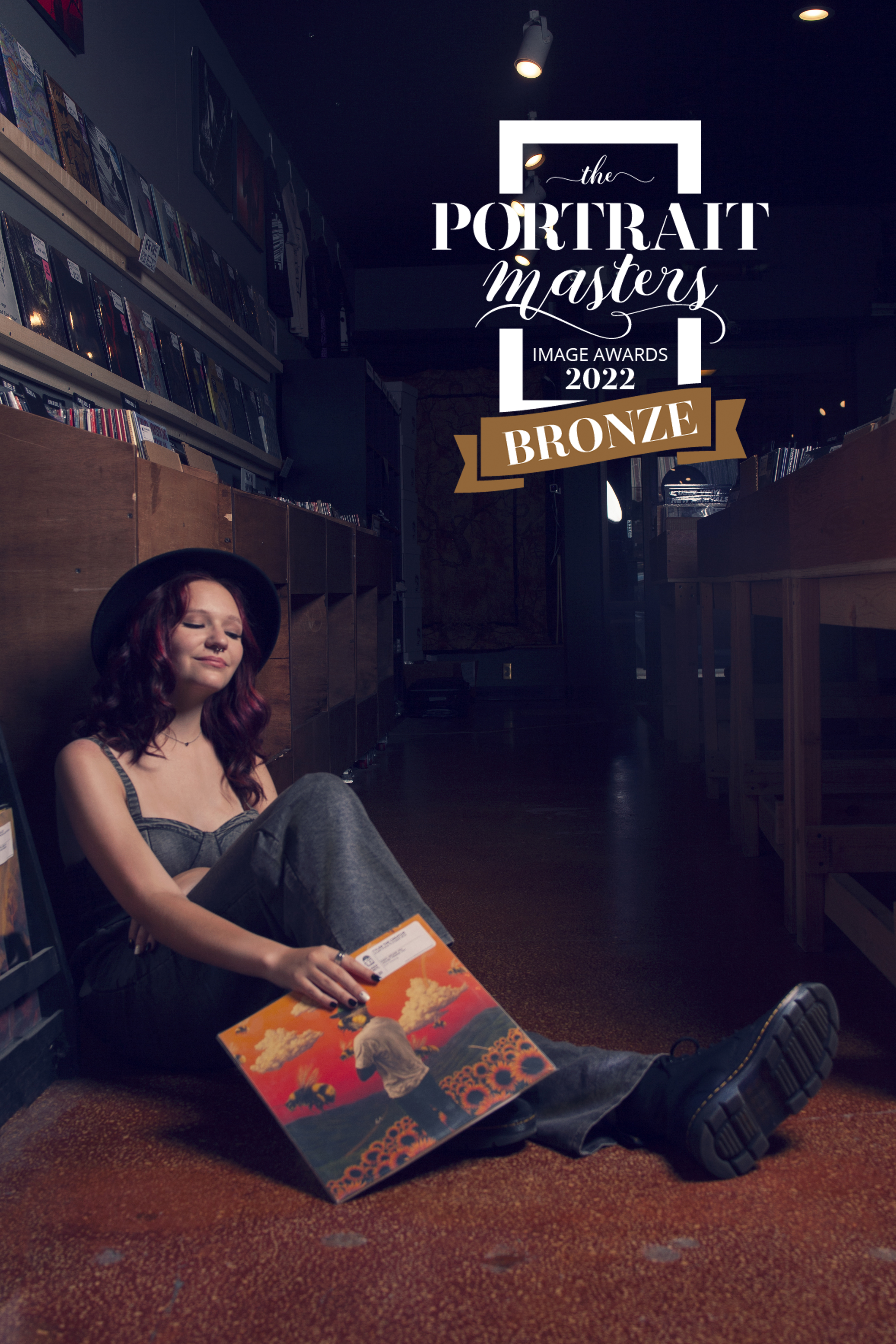 A person with red hair sitting on the floor of a record store, holding a colorful album cover, with shelves of records in the background. The image includes a banner indicating a Bronze award from the Portrait Masters Image Awards 2022.