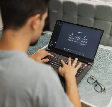 A college student using a laptop with the ChatGPT interface open, working attentively on a project. The laptop screen displays the ChatGPT application, highlighting features relevant to student needs. The text accompanying the image reads, "ChatGPT will be free for college students through May," emphasizing access to AI tools for educational purposes. A pair of glasses lies on the table beside the laptop, suggesting a study environment.