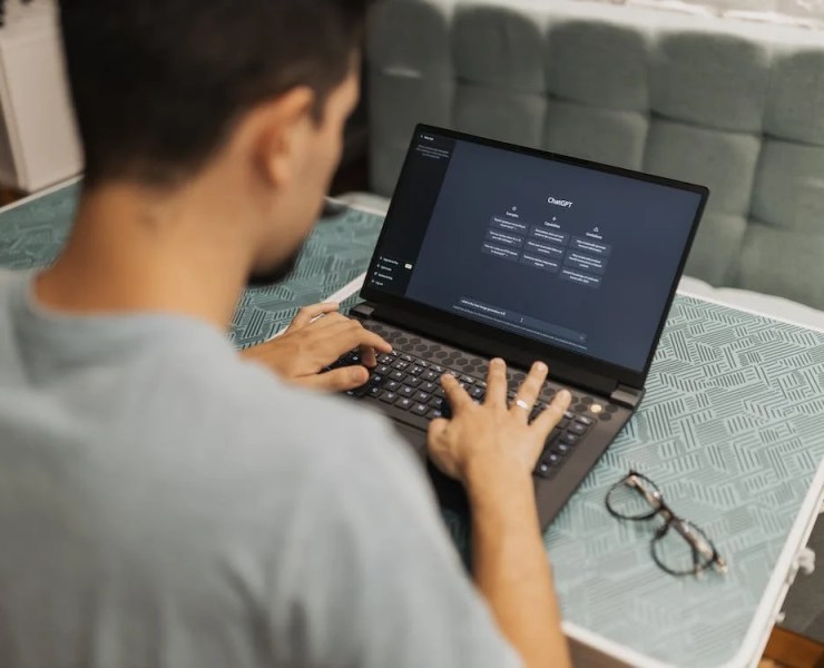 A college student using a laptop with the ChatGPT interface open, working attentively on a project. The laptop screen displays the ChatGPT application, highlighting features relevant to student needs. The text accompanying the image reads, "ChatGPT will be free for college students through May," emphasizing access to AI tools for educational purposes. A pair of glasses lies on the table beside the laptop, suggesting a study environment.