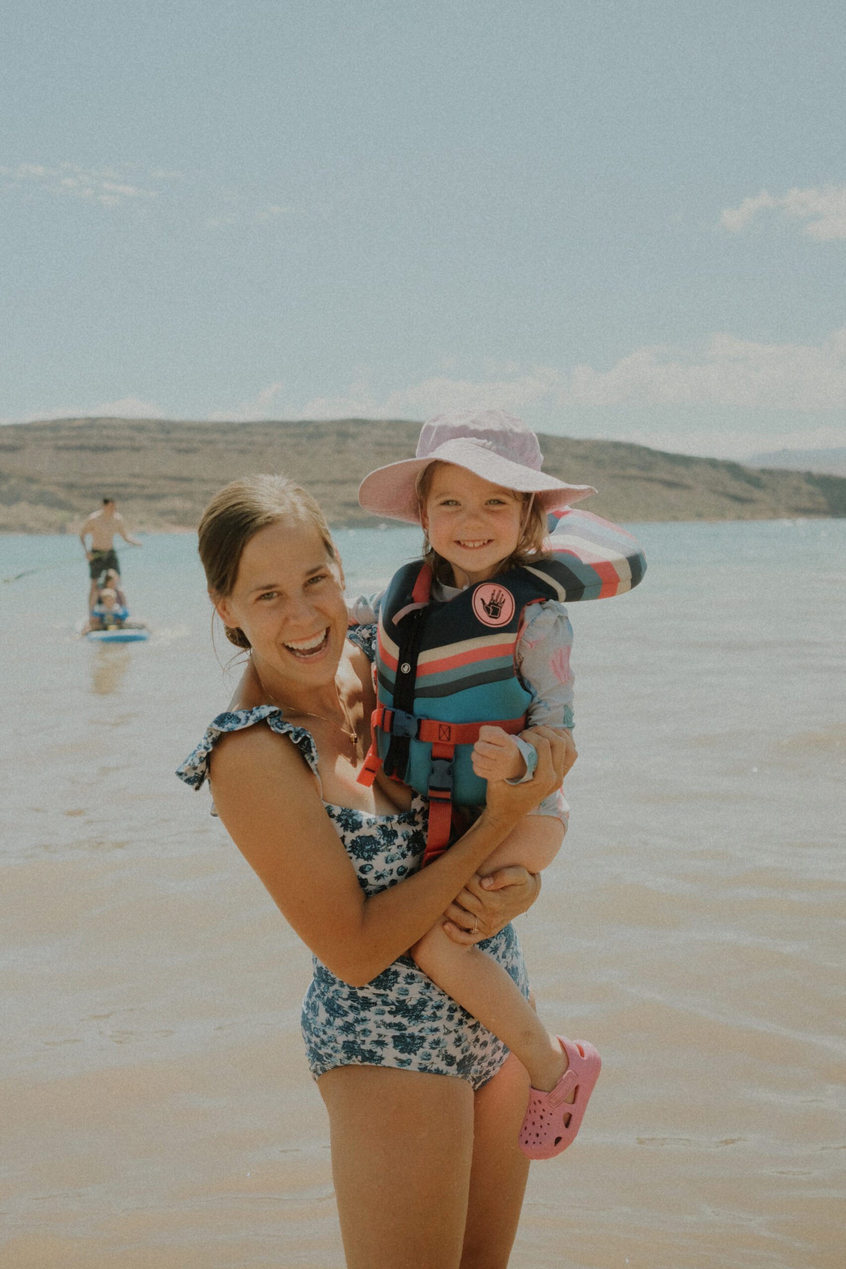 Mom holding her toddler in a life jacket on a sunny lake shore