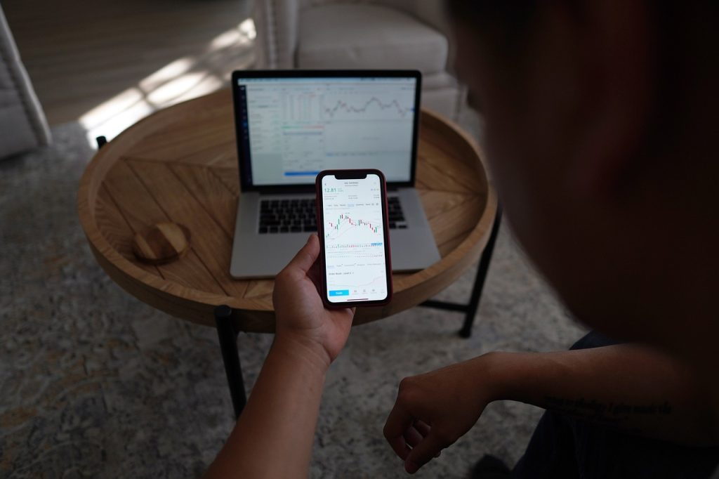 holding a phone with a trading chart and a laptop on top of a table in the back