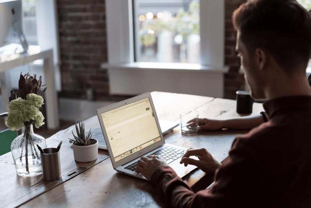 A man in a burgundy shirt working at a laptop on a wooden desk, surrounded by plants in a bright, modern office with an exposed brick wall.