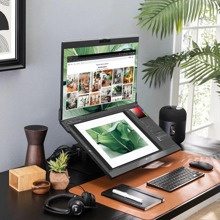 Dual-screen laptop angled on a home office desk showing photo editing software, accompanied by a keyboard, mouse, and plants.