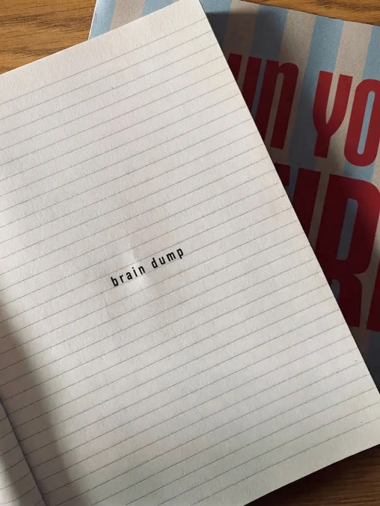 An open lined notebook page with the words 'brain dump' centered on the page in black type, resting on a wooden table next to a second book. solo rituals