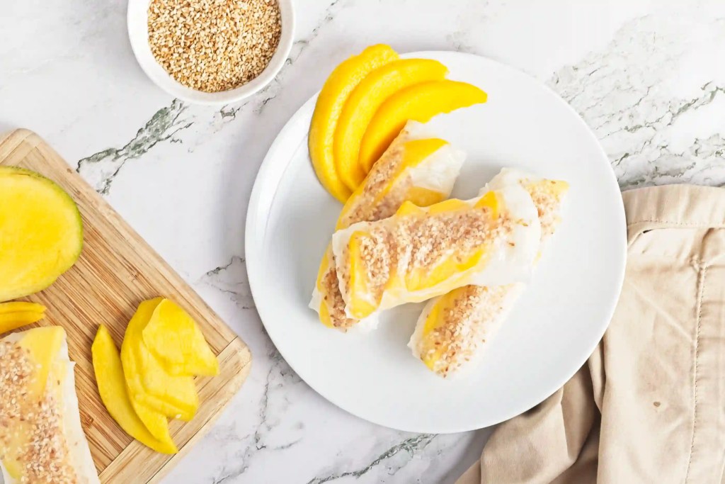 vegan and gluten free mango sticky rice spring rolls on a wooden cutting board next to half a mango