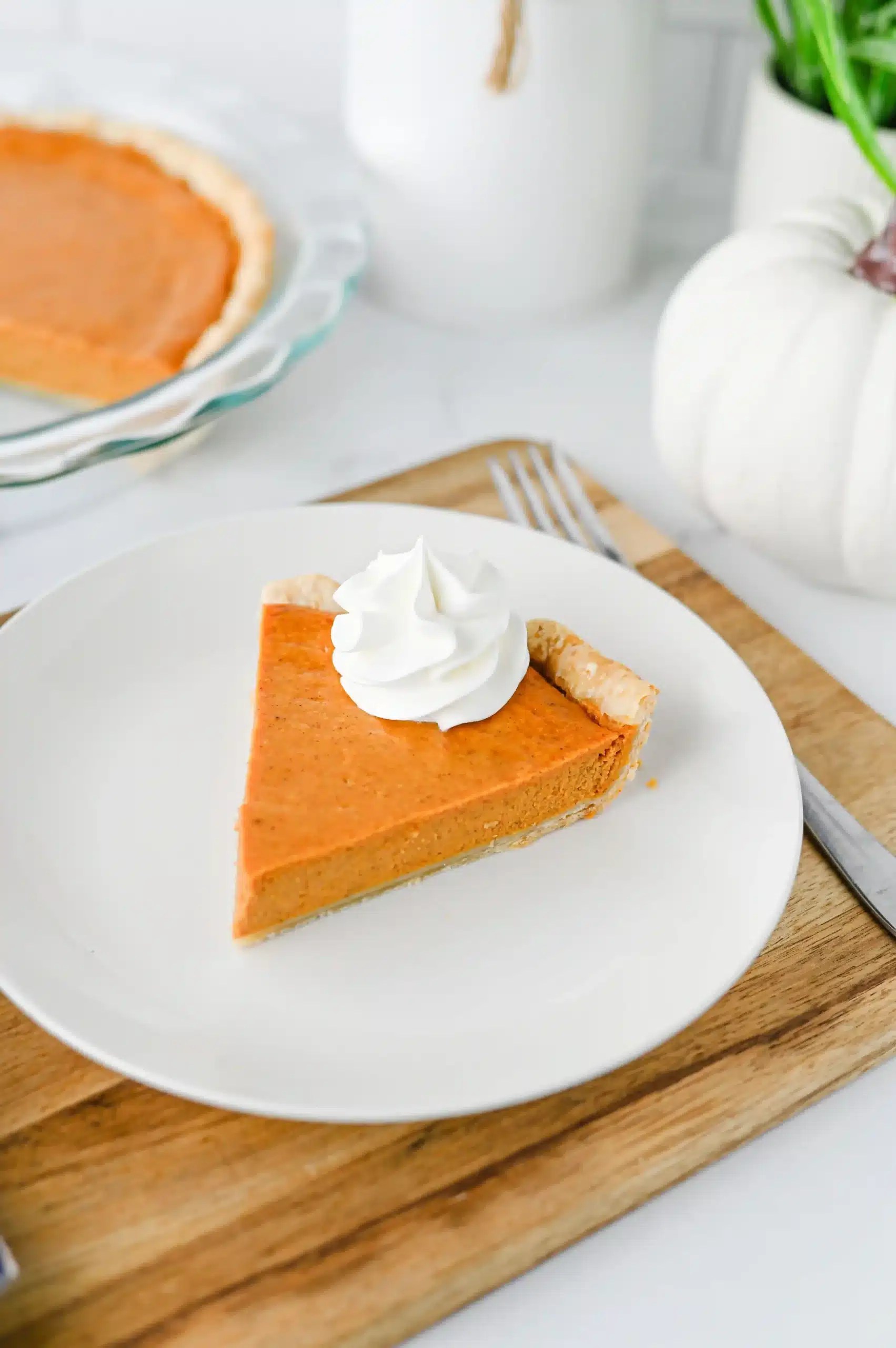 a slice of gluten free pumpkin pie with a dollop of whipped cream on a white plate resting on a wooden board with a fork