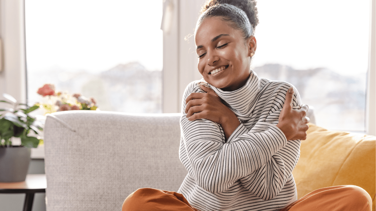 A young woman sitting comfortably at home, hugging herself and smiling contentedly, embodies the essence of self-love and personal acceptance.