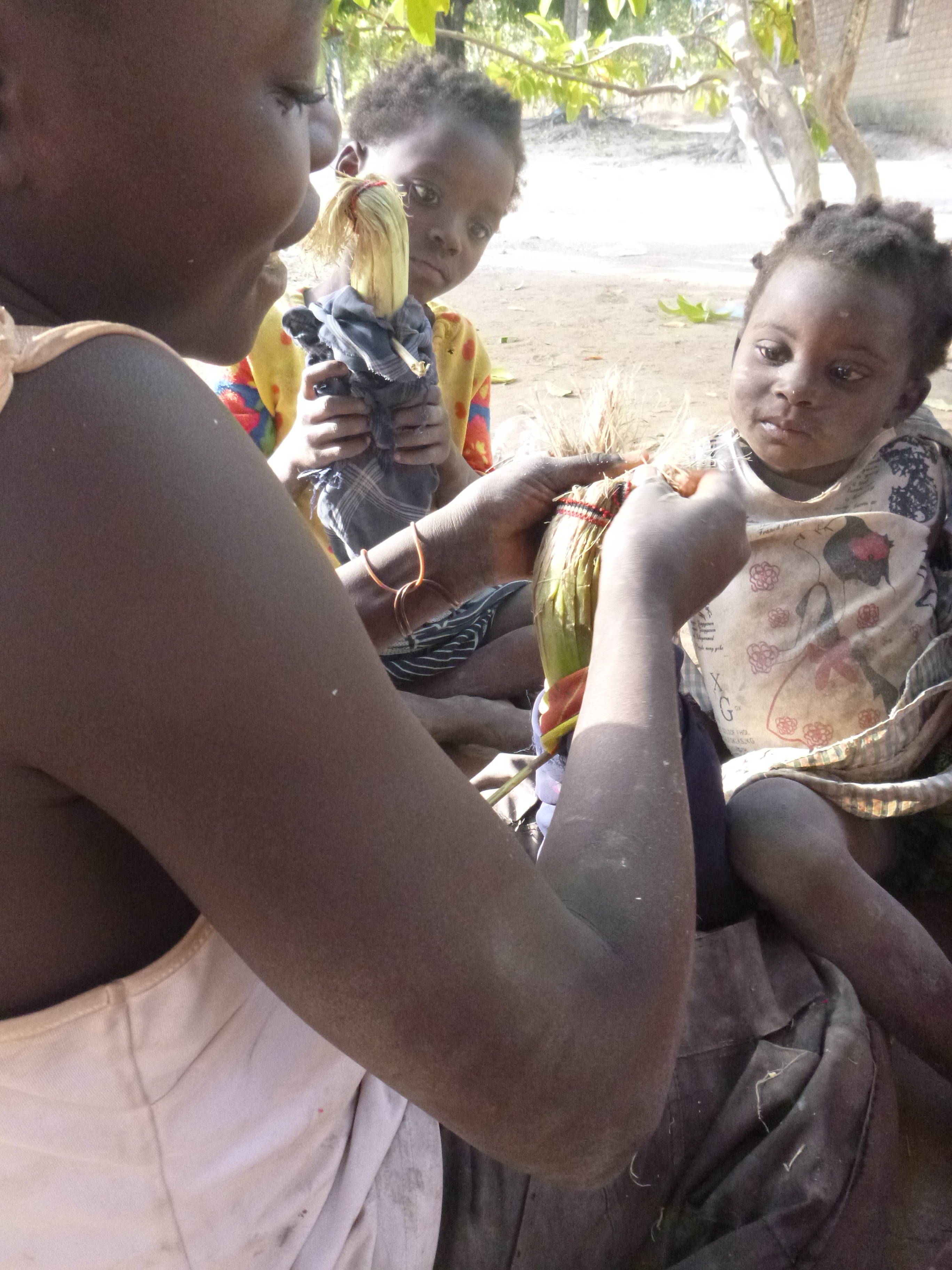 Doris helps Minus braid her doll's hair while Jess watches with her own doll. The dolls are made from banana stalks.