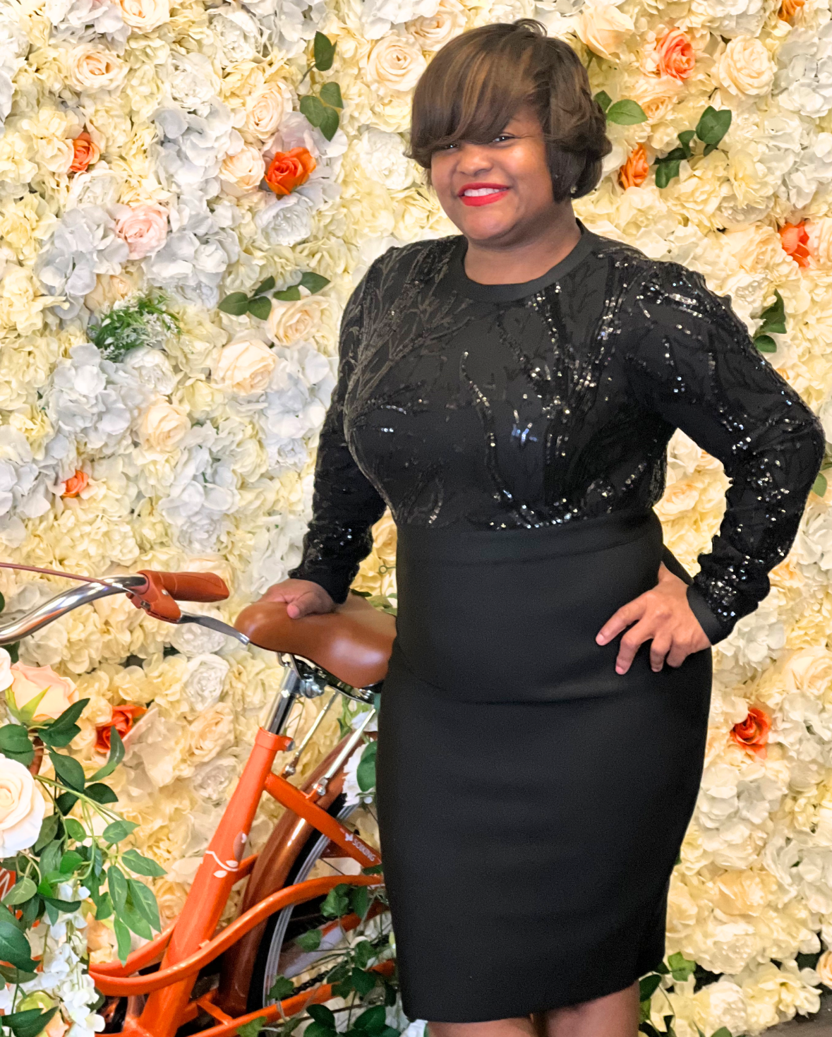 Woman in elegant black dress poses with orange bicycle against floral wall.