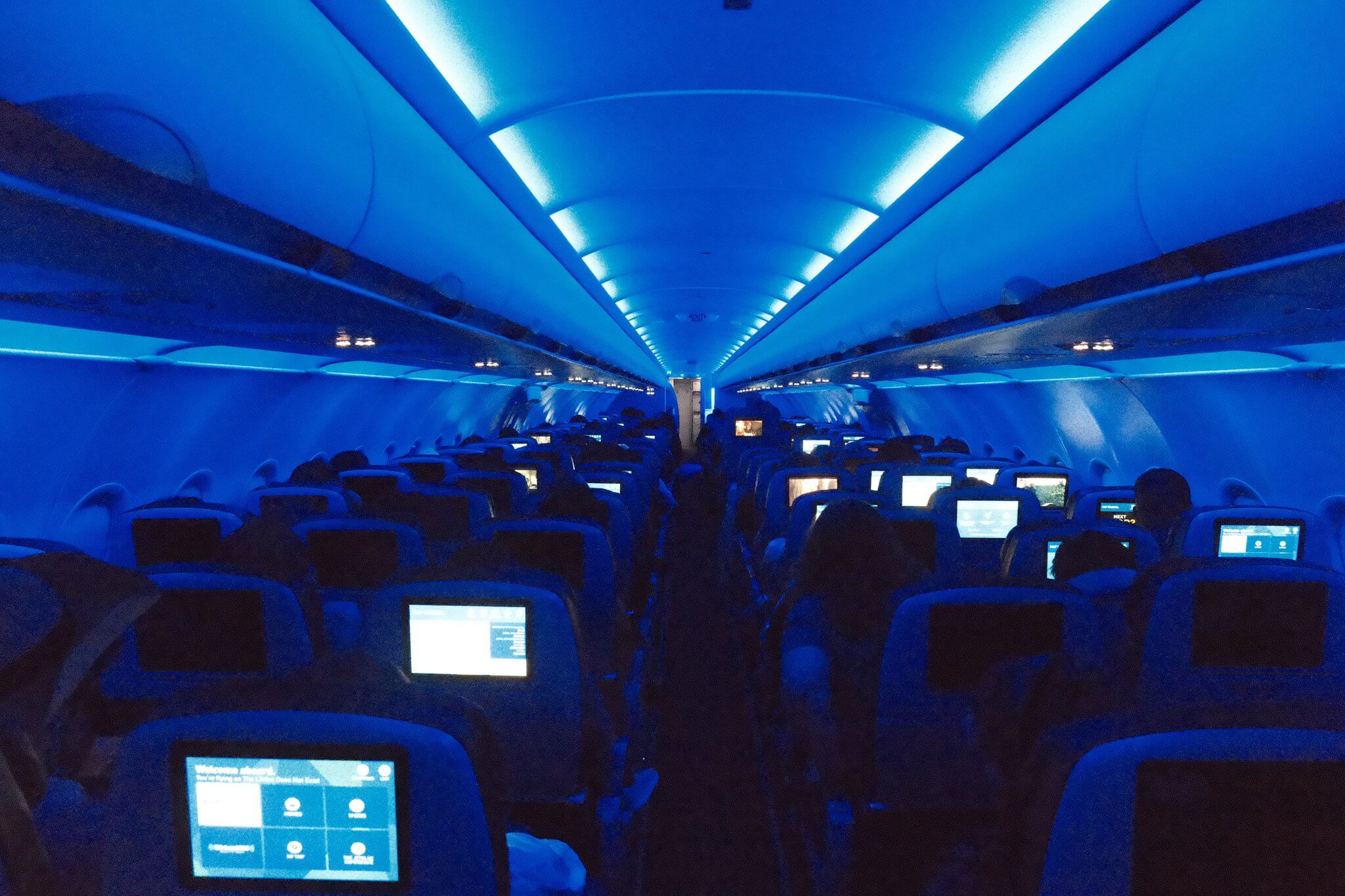 Interior of a modern aircraft cabin bathed in vivid blue mood lighting; rows of seats recede toward the front of the plane, seatback entertainment screens glow in the darkness, and overhead bins are lit from within — likely a JetBlue or similar carrier on a long-haul night flight
