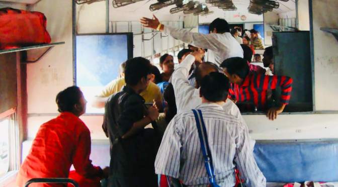 Crowded interior of an Indian Railways unreserved carriage; passengers in brightly coloured clothes stand packed together in the aisle, one man gestures overhead, overhead luggage racks and ceiling fans are visible — India