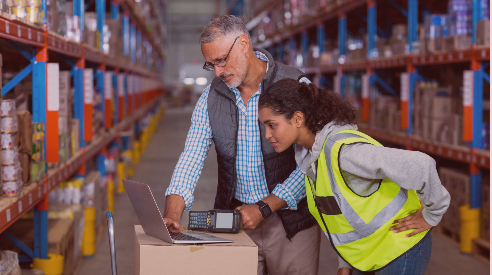 Two warehouse workers reviewing information on a laptop while standing among tall shelves stocked with inventory, one holding a barcode scanner.