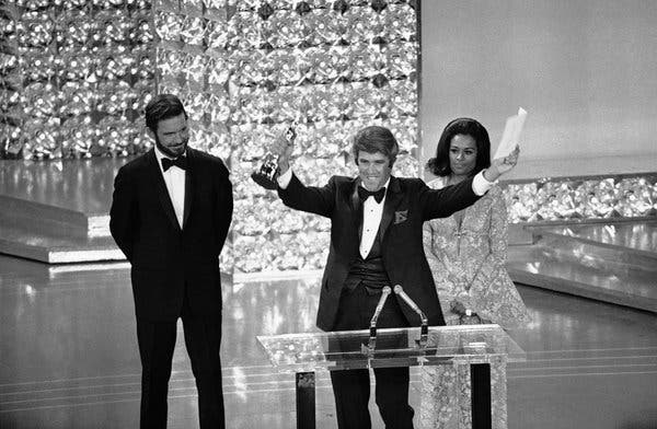 Mr. Bacharach, wearing a tuxedo and smiling, holding up both arms in triumph, a statuette in his right hand, at the Oscar podium.