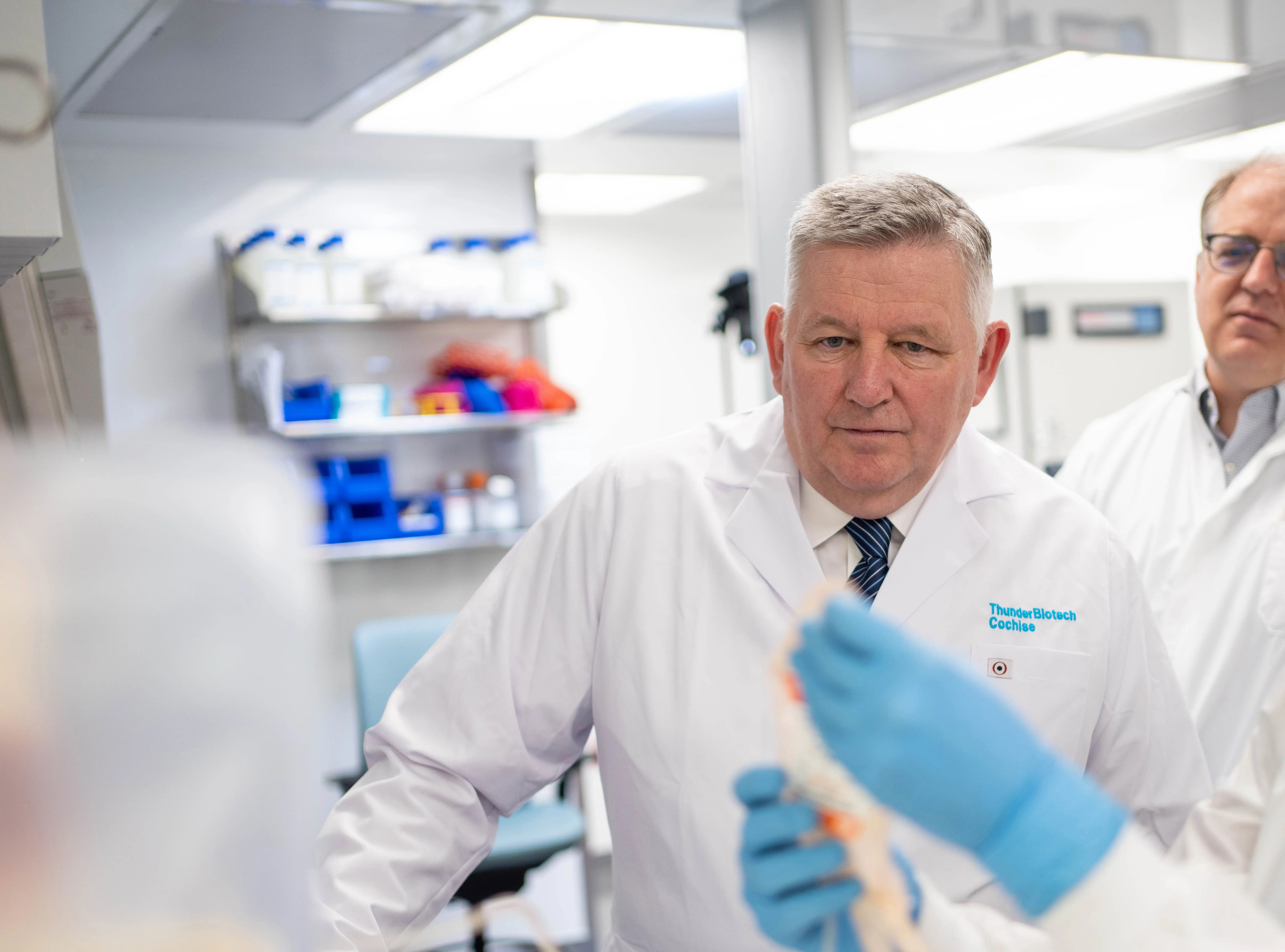 Dr. Kim O’Neill and Dr. Kevin Gurney looking on as a lab member shows them a sample in a test tube.