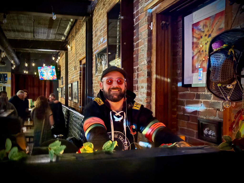 A cheerful bartender wearing sunglasses and a hoodie smiles while standing behind a bar, with a rustic brick wall in the background and dim lighting.