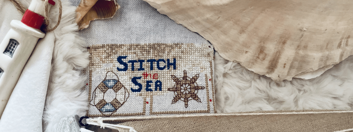 Third section of the Stitch the Sea SAL featuring a title, Stitch the Sea, in blue surrounded by sparkling sand and a ship's wheel and life preserver.