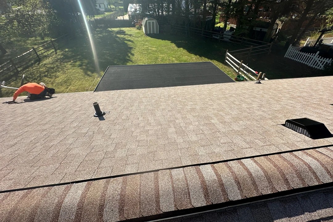 Freshly installed tan shingle roof with ridge caps and vents, shown from above as a worker in an orange shirt finishes detail work near the edge.
