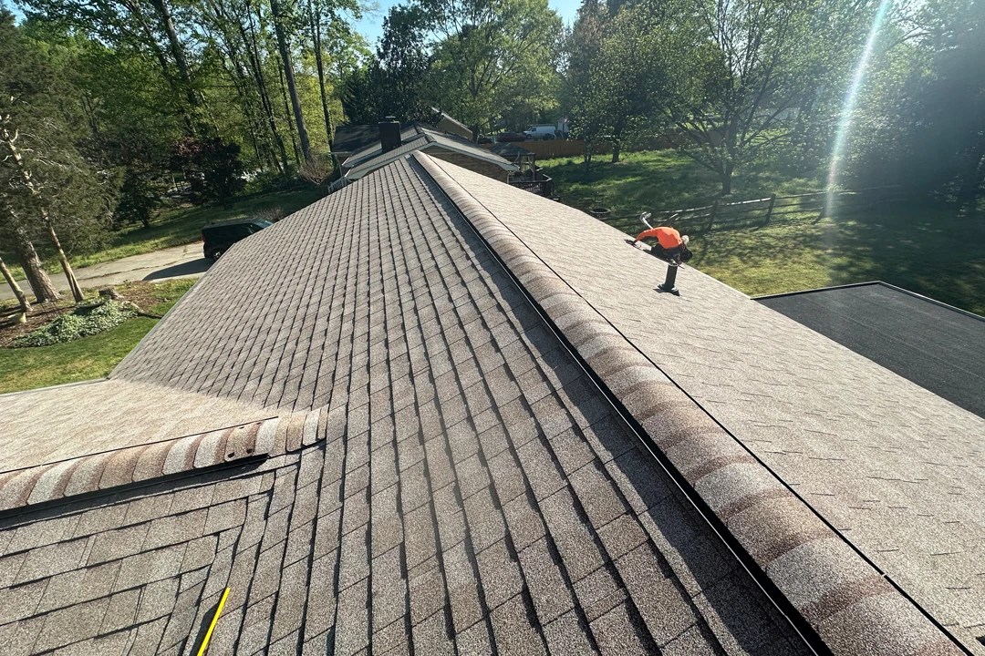 Newly installed tan asphalt shingle roof with ridge caps, viewed from above as a worker in an orange shirt completes finishing touches.