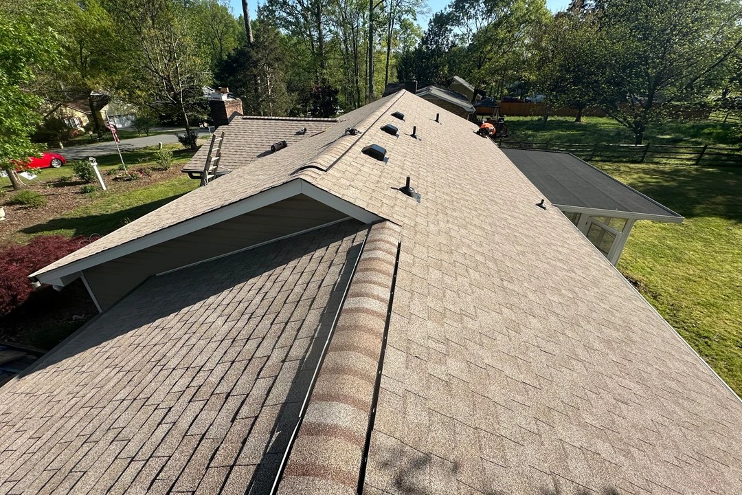 New tan asphalt shingle roof with ridge caps and multiple vent installations, shown from above on a sunny day.