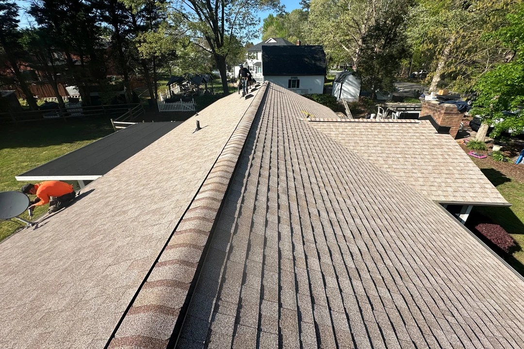 Freshly installed tan asphalt shingle roof with ridge caps, shown from above as workers finish details near the edges on a sunny day.