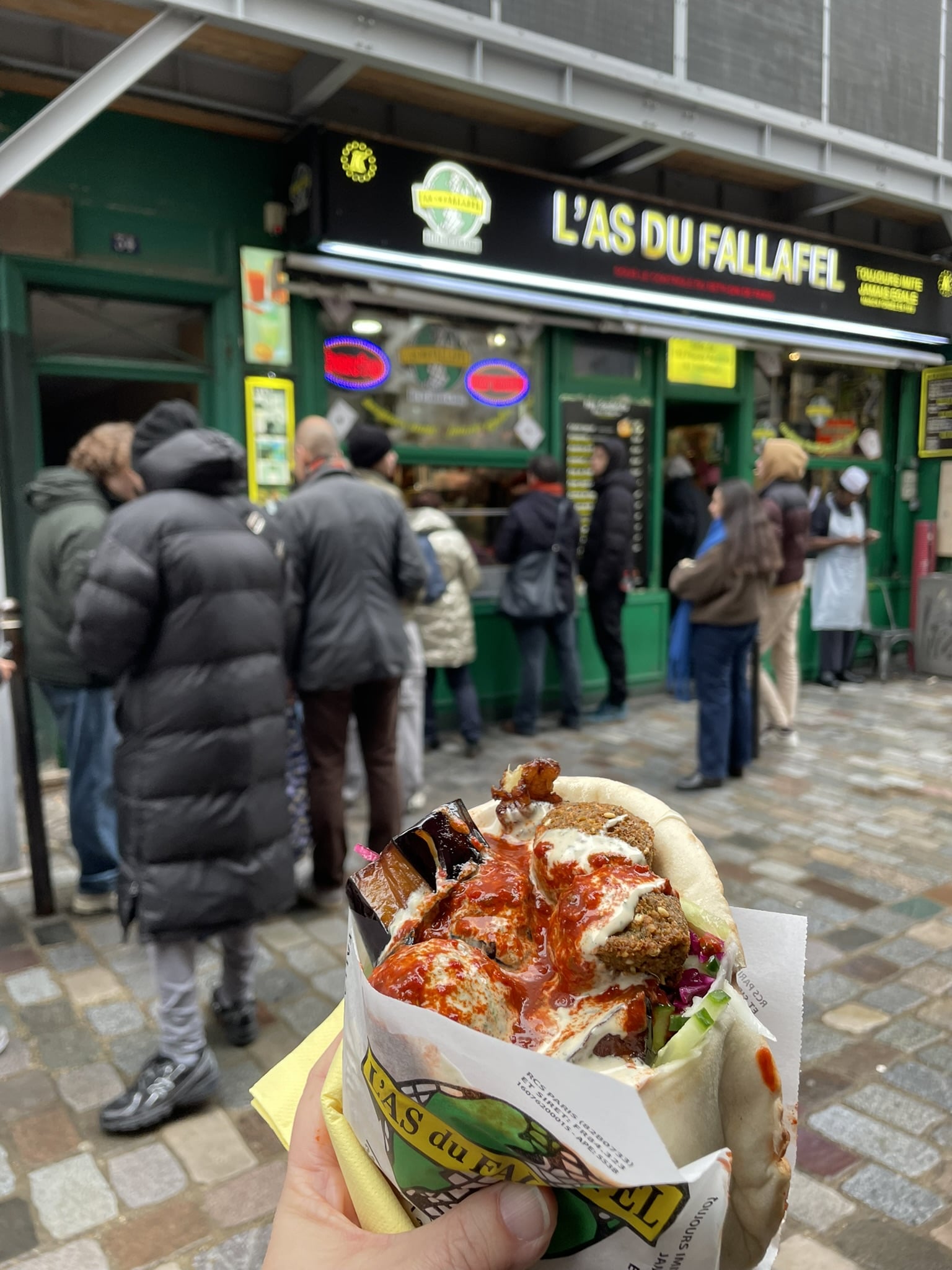 Hand holding a large falafel sandwich with the L'As du Fallafel storefront in the background
