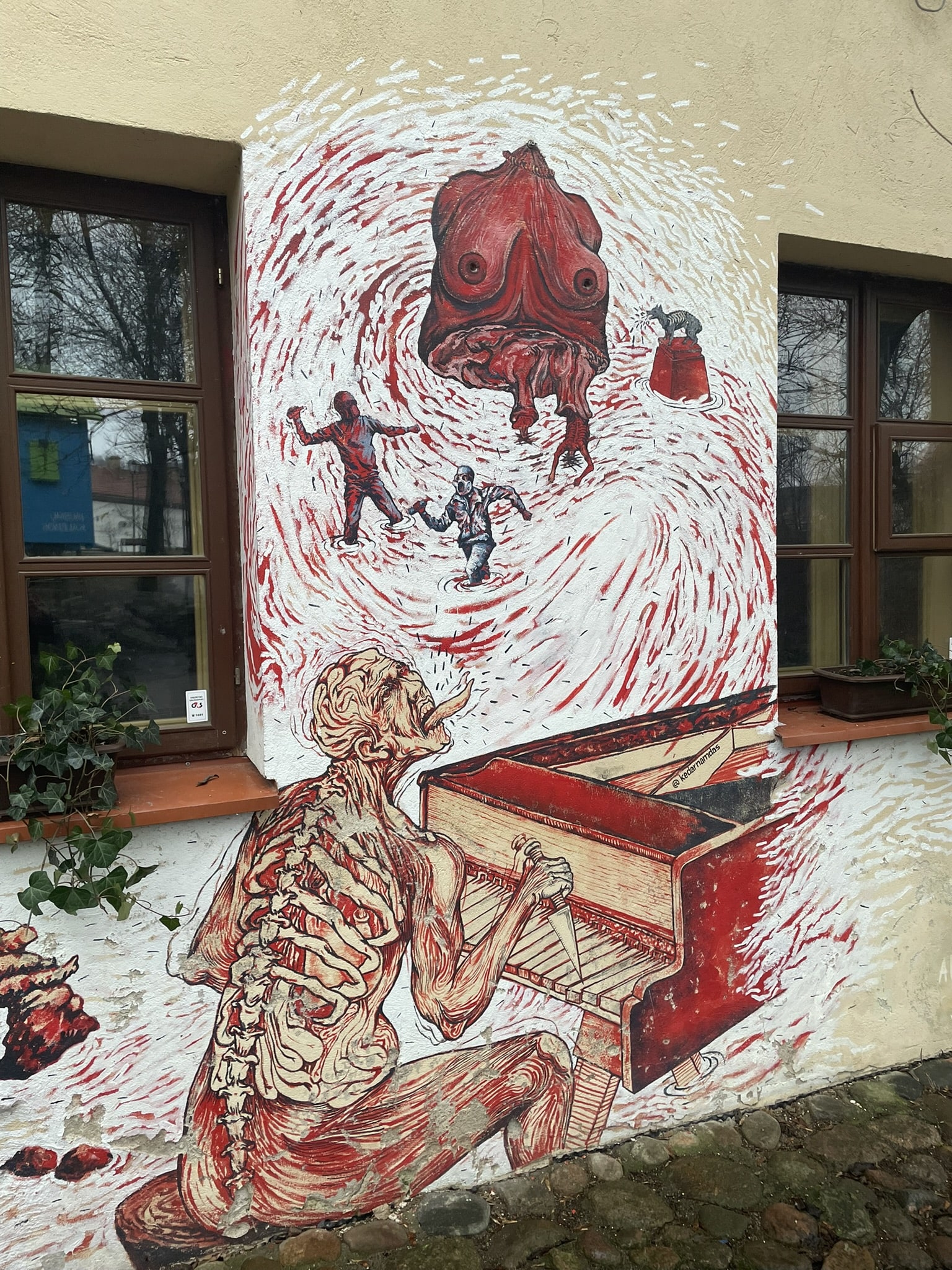 street art painted on a building featuring a man with exposed bones and circulatory system playing a piano while above him is a scene of red and white fantasy? Featuring a woman's bare breasted torso and two small male figures