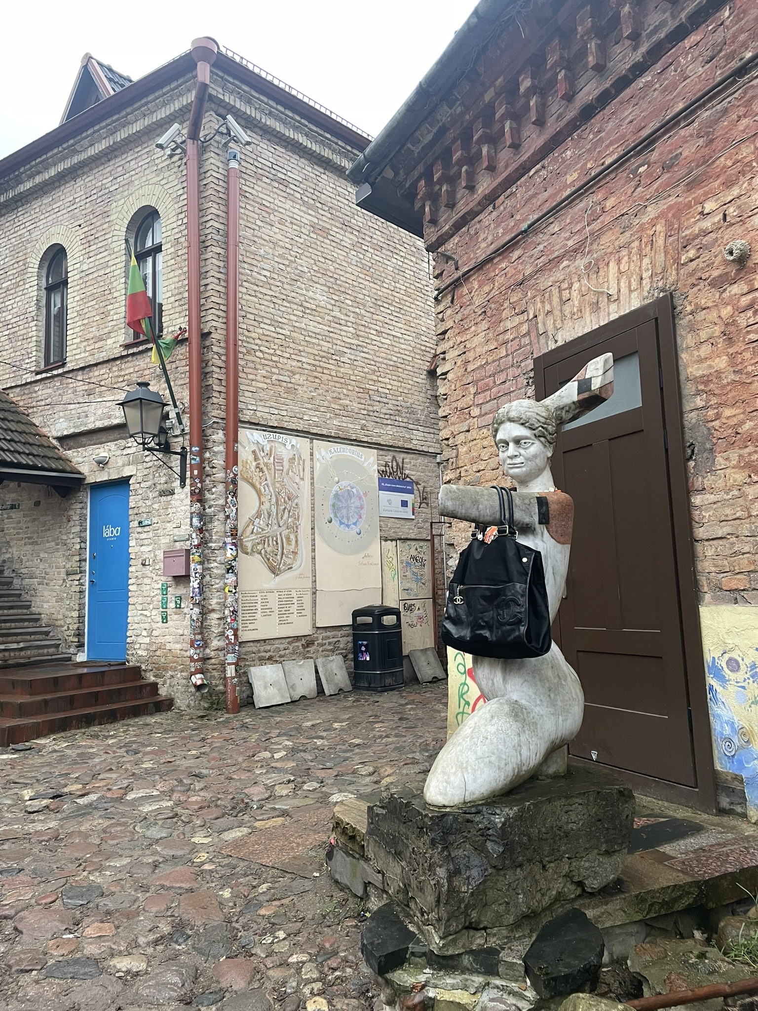 brick building and cobblestone street with some kind of statue/mannequin on a rough stone pedestal. One arm is outstretched but broken off at the elbow and a purse is hung on the arm