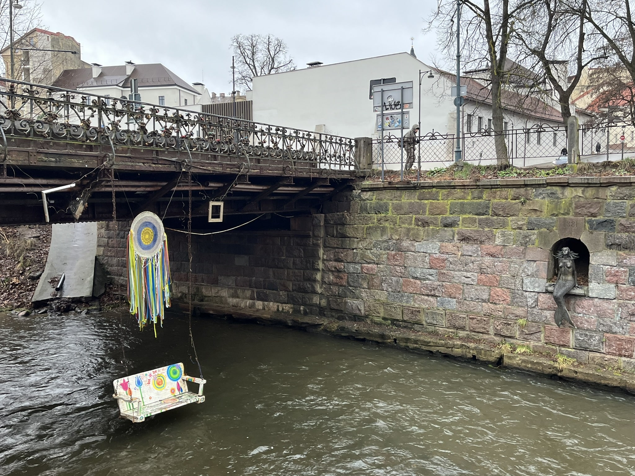Bridge over a river. Inset into the wall on one side of the river is a statue of a mermaid. A porch swing hangs off the bridge into the middle of the river.