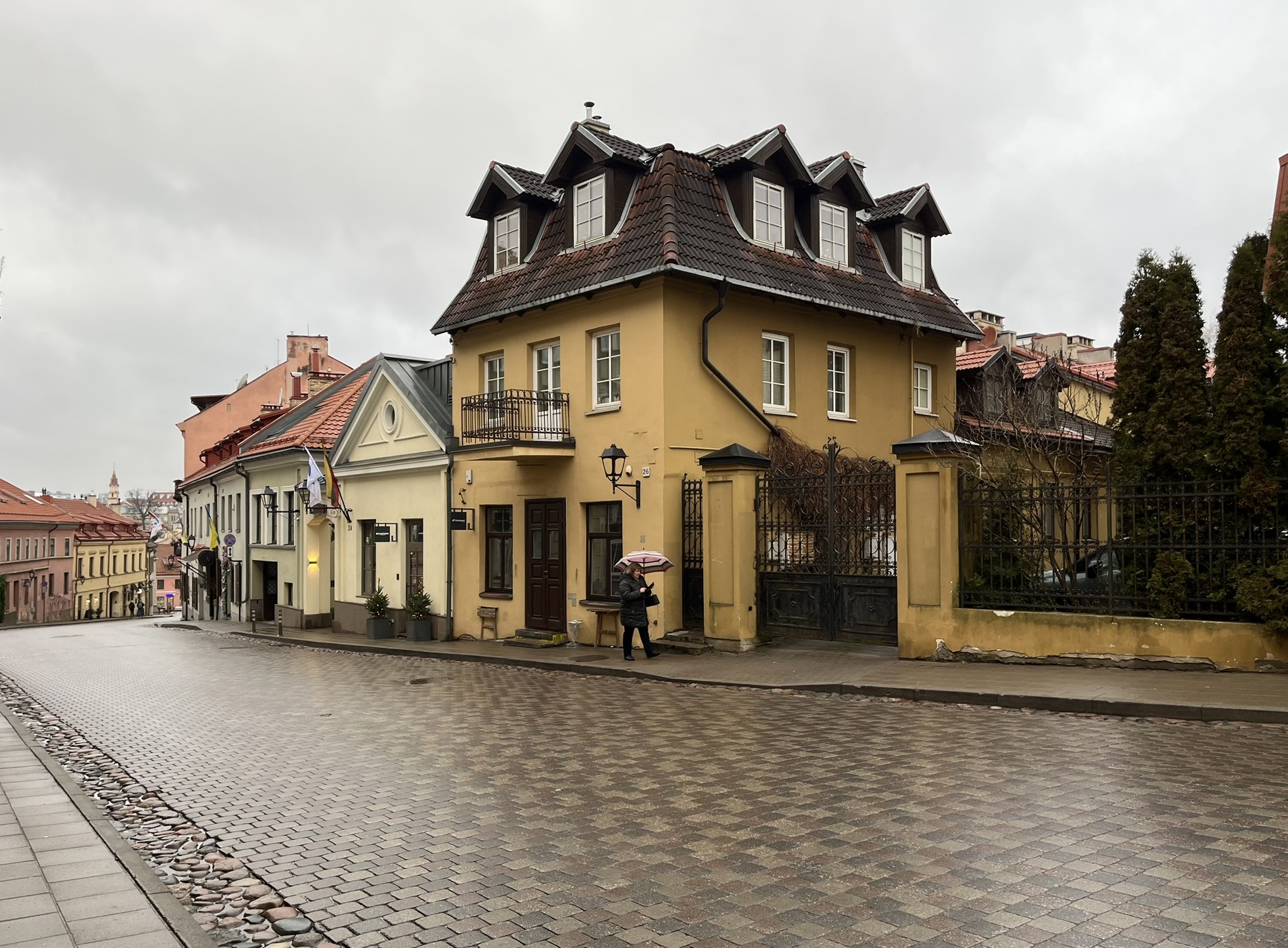 slightly inclined brick street featuring pastel buildings and a woman walking by with an umbrella