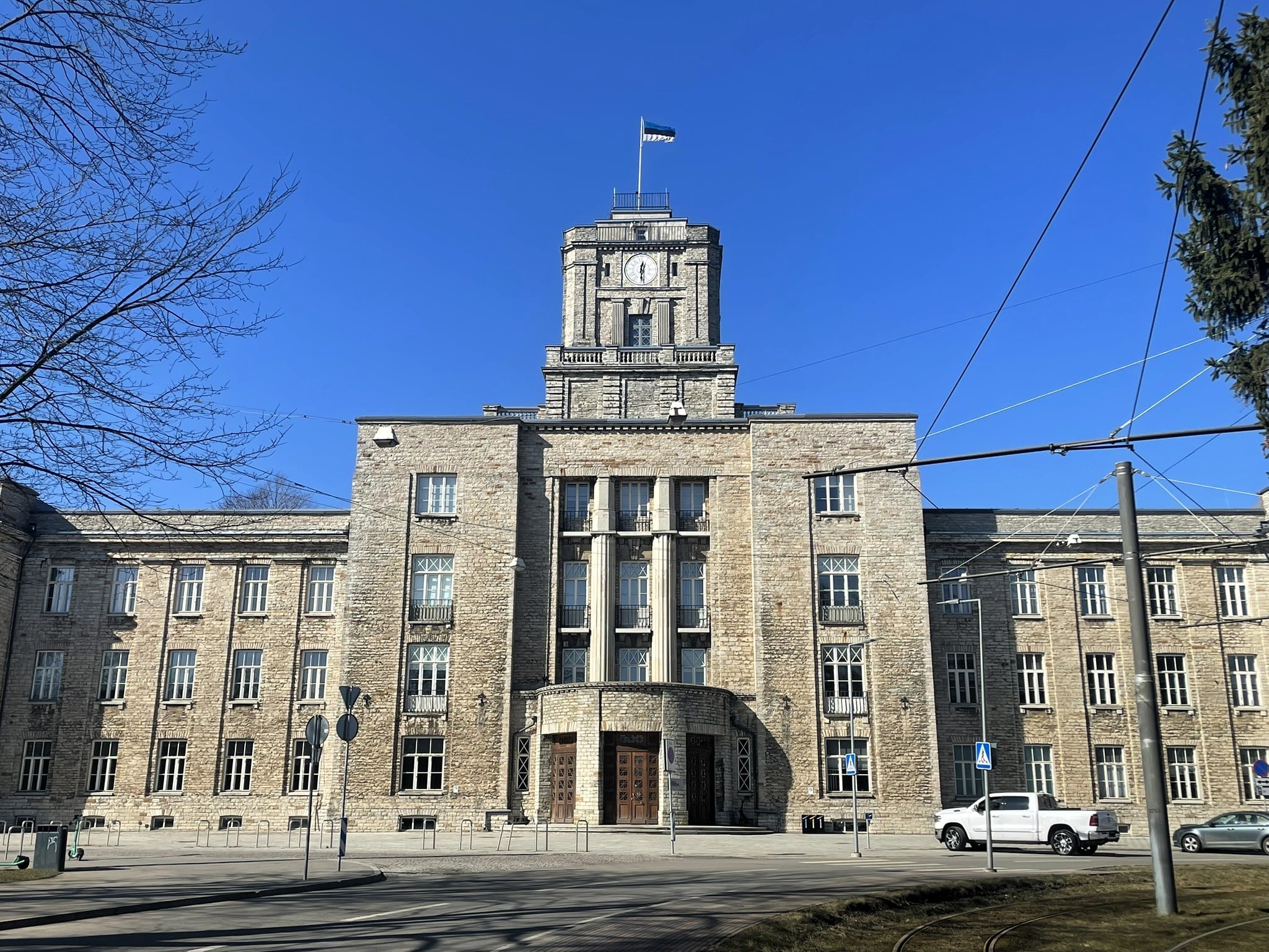 Imposing limetstone building with a clock tower, pillars, and curved entrance. Topped with an Estonian flag.