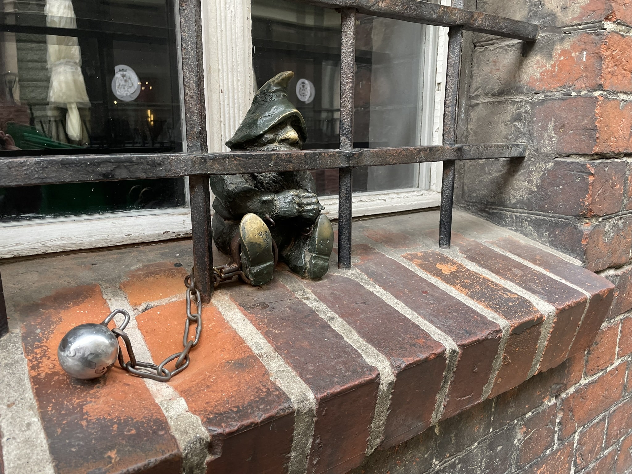 small statue of a gnome with a ball and chain attached, sitting in a barred window well