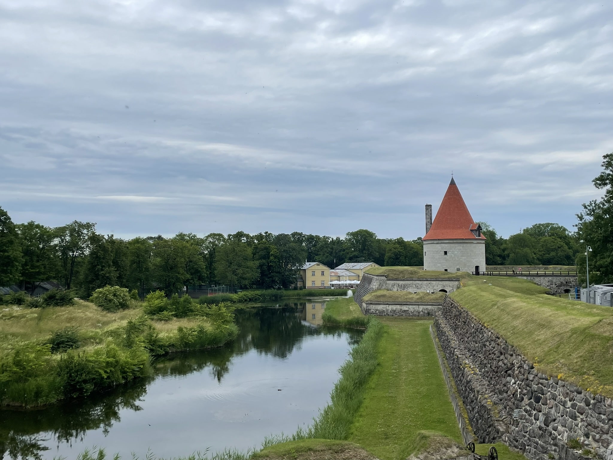 A moat and some turf and stone castle fortifications, with a conical tower in the distance