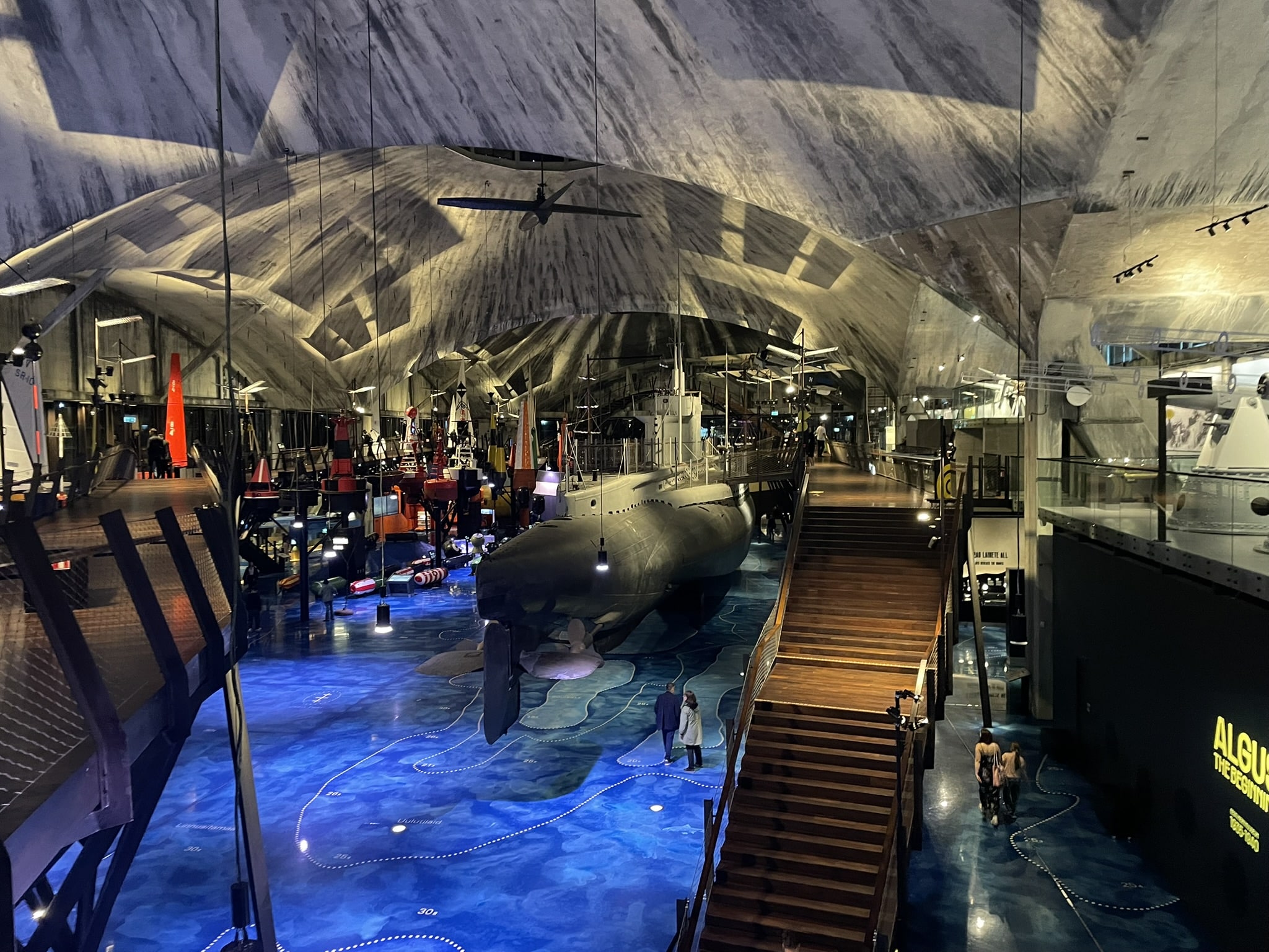 Cavernous space with a blue floor, grey walls and ceiling, and several maritime items displayed