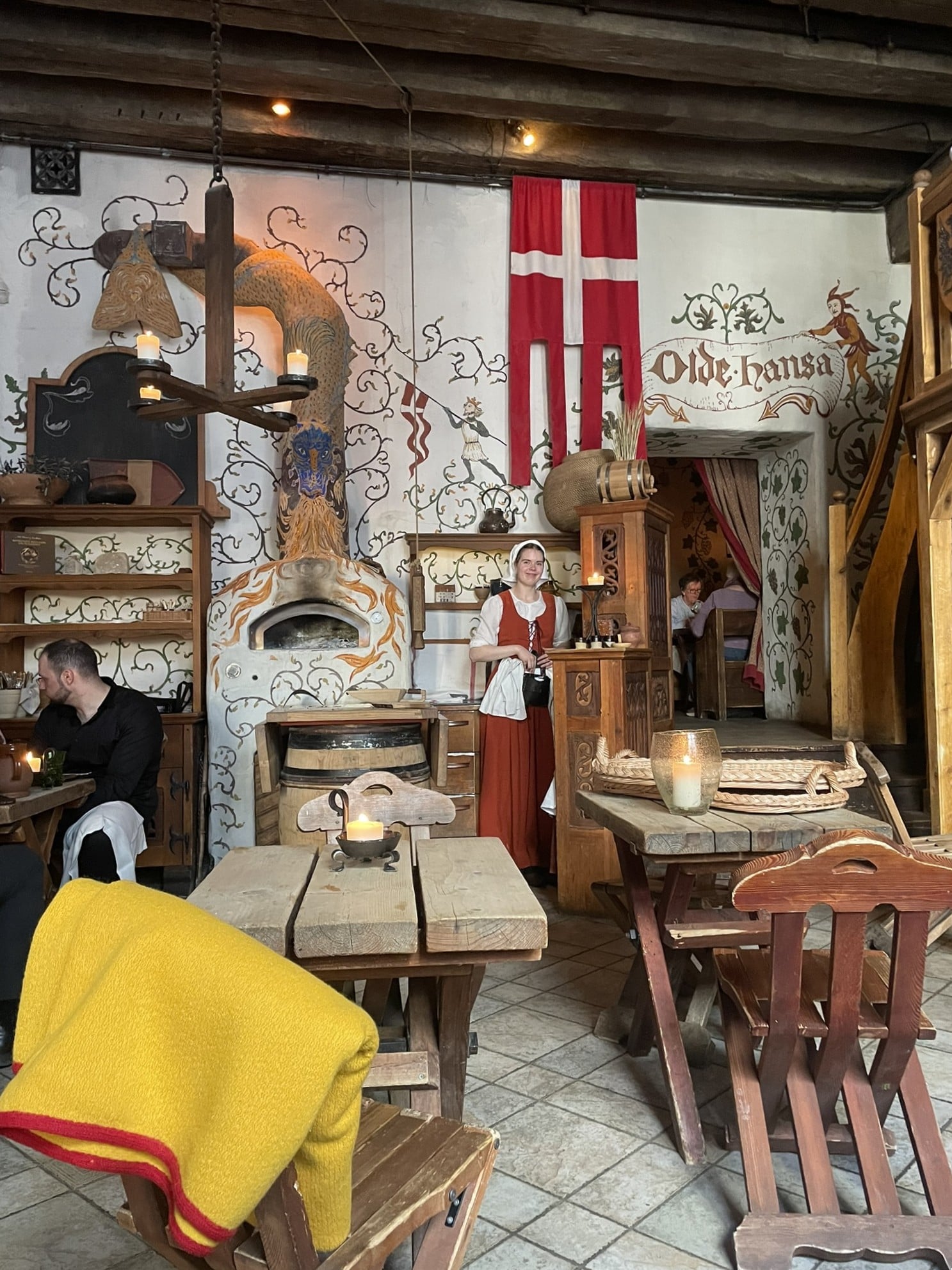 A woman in medieval costume standing in a dining room of wooden furniture.