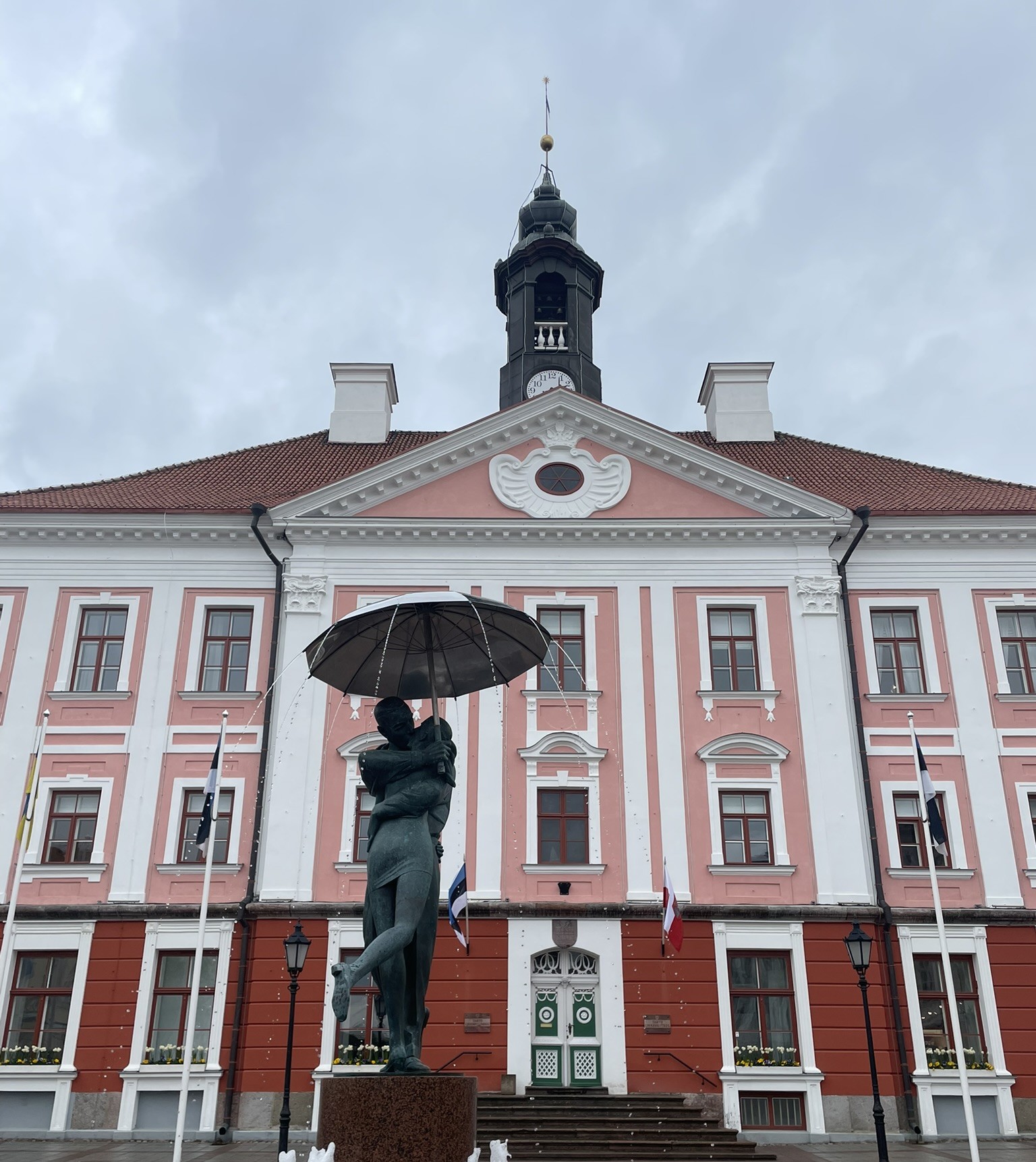 pink baroque building with a tower, with a statue of two people embracing under an umbrella in front.