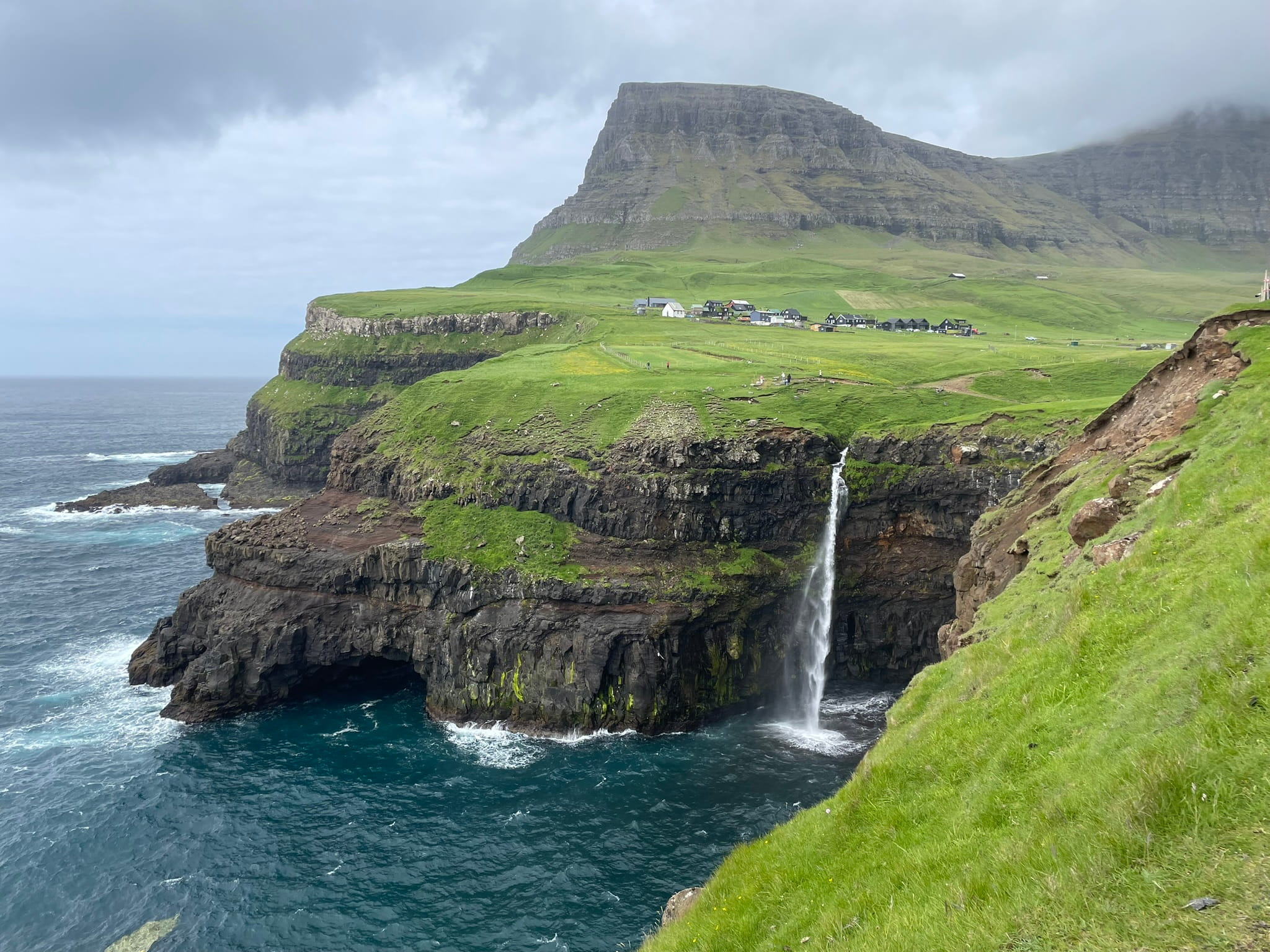 Green mountain in the distance, with the foreground showing a waterfall emoytying directly off of a cliff and into the ocean