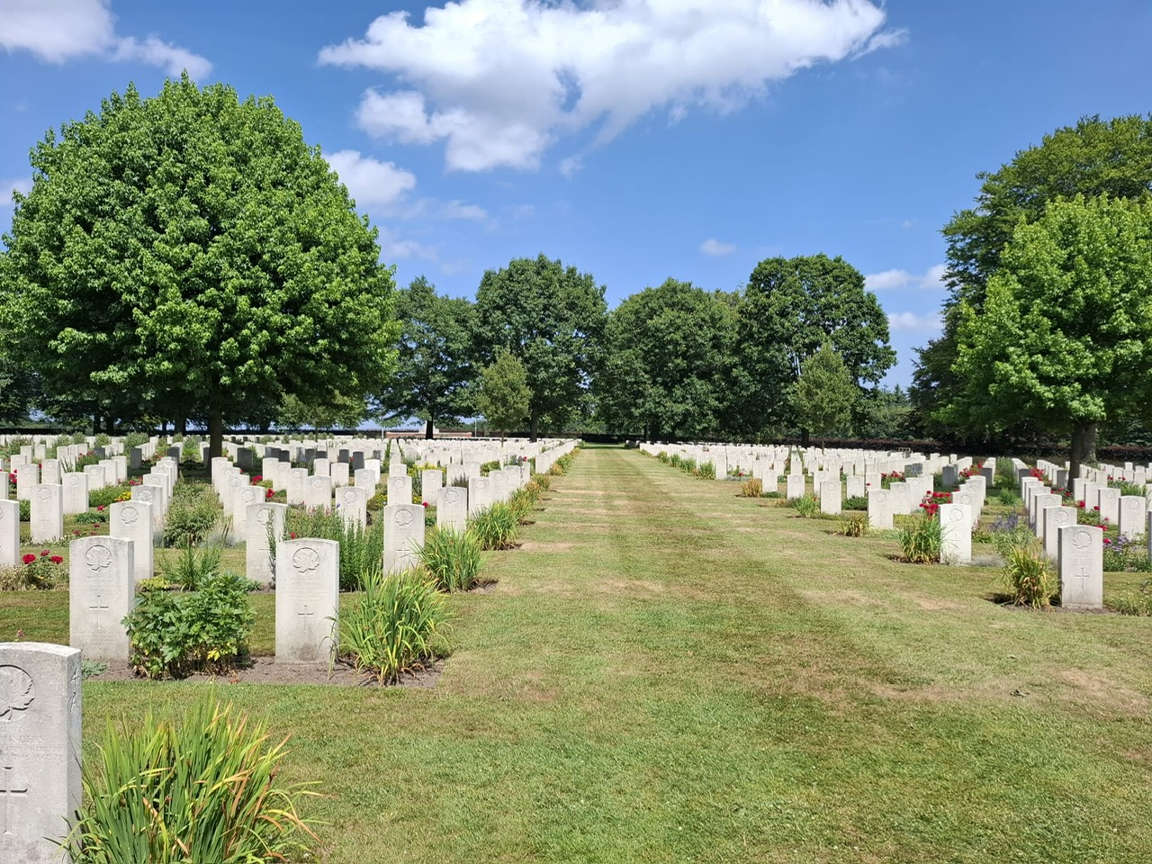 Foto van de Canadese militaire begraafplaats in Groesbeek. Rijen identieke witte grafstenen staan netjes in rechte lijnen op een goed onderhouden grasveld. Tussen de grafstenen zijn kleine tuintjes met groene planten en wat bloemen aangelegd. De begraafplaats is omringd door grote, volle groene bomen die een natuurlijke omlijsting vormen. Er loopt een gemaaid graspad over het midden van de begraafplaats. De lucht is helderblauw met enkele witte wolken. De scene straalt rust, waardigheid en respect uit voor de gevallenen die hier begraven liggen. Het onderhoud van de begraafplaats lijkt zeer zorgvuldig te gebeuren, wat te zien is aan het verzorgde gras, de nette plantvakken en de schone, rechte grafstenen. Dit type begraafplaats is kenmerkend voor de Commonwealth War Graves Commission, die oorlogsbegraafplaatsen onderhoudt ter nagedachtenis aan de militairen die zijn gesneuveld tijdens de wereldoorlogen.