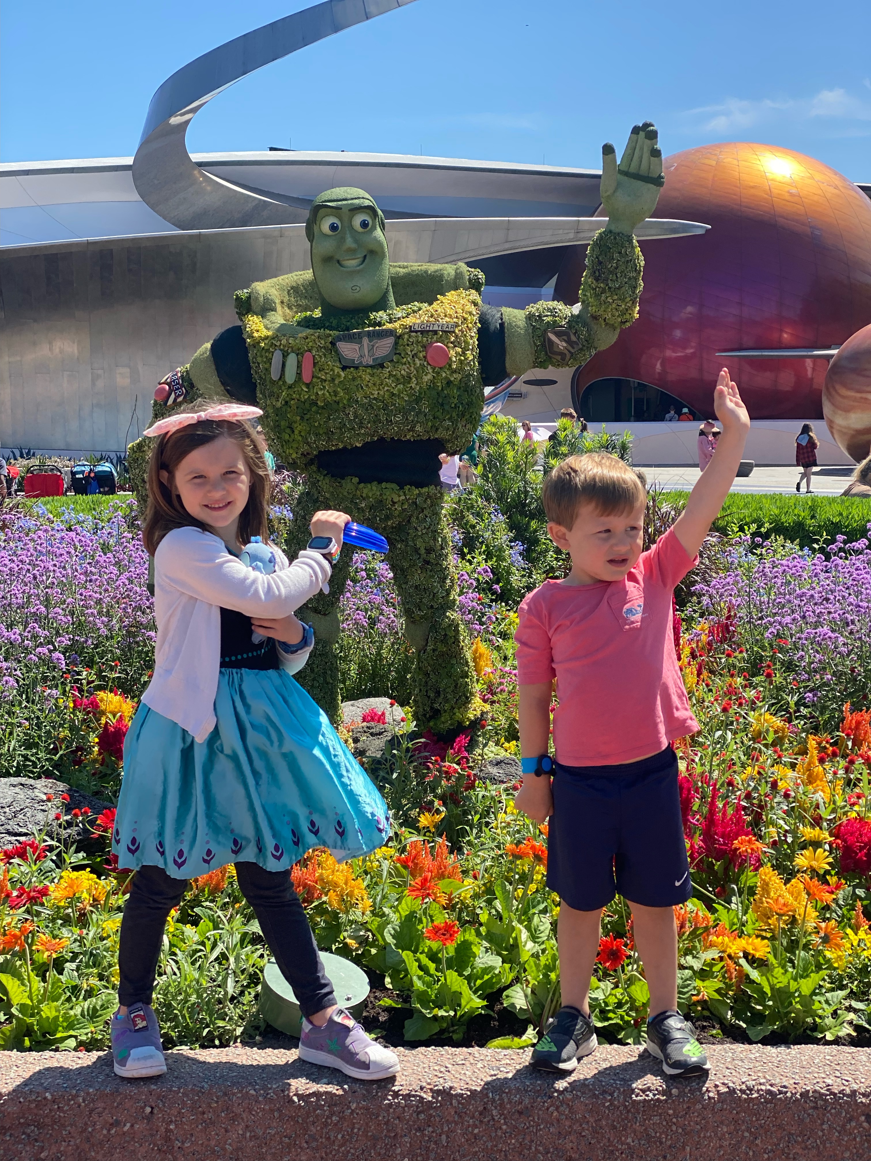 An image of two children posing in front of a large, green, anthropomorphic figure. They are in front of a flower garden a...