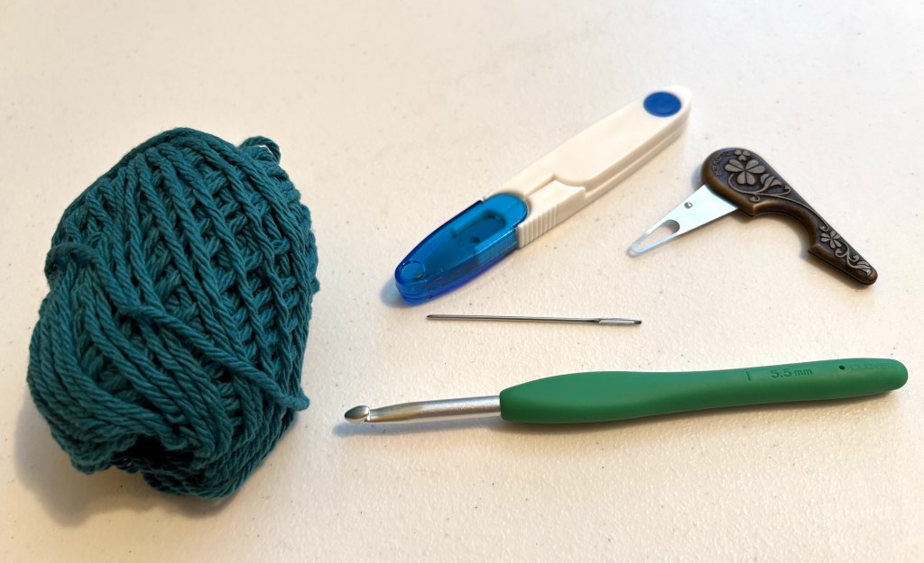 blue yarn, a crochet hook, a needle, a yarn threader, and scissors sitting on a table.