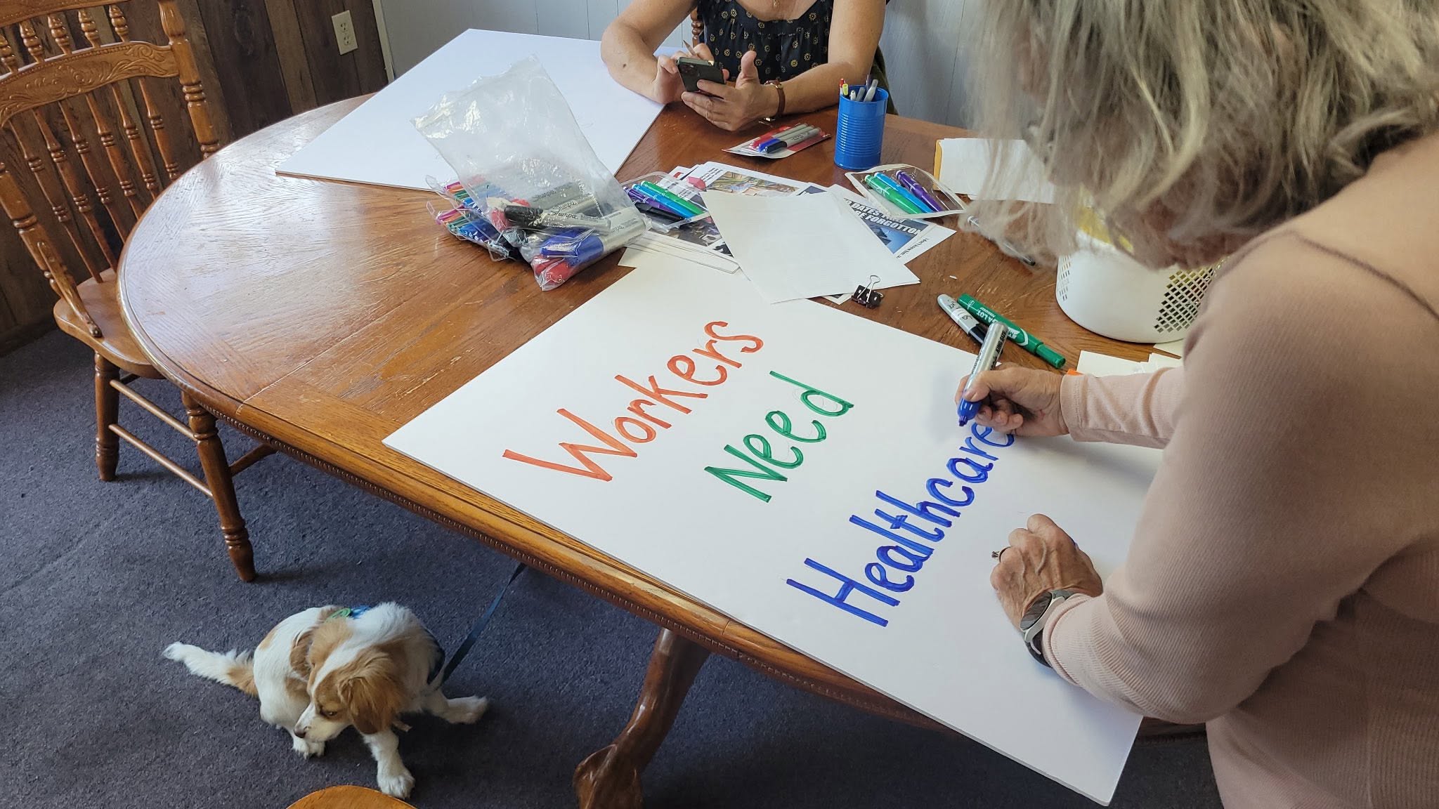 Two people sit at a wooden table crafting a large colorful poster; a small dog lounges at the carpeted floor nearby.