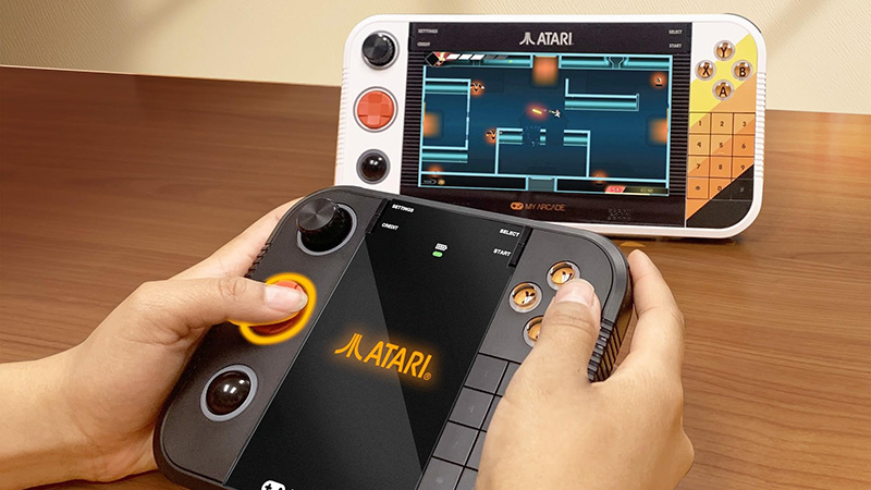 Two Atari Gamestation Gamepads, one in use and another showcasing its retro design and keypad, on a wooden surface.