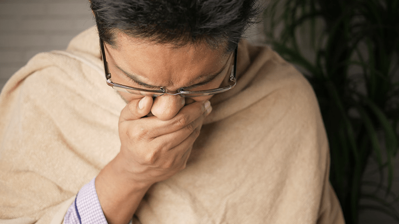 A man wrapped in a beige blanket, covering his mouth, symbolizing flu symptoms and illness.